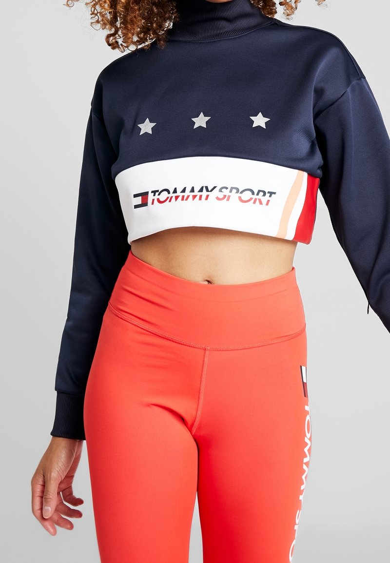 Femme portant un sweat-shirt court bleu marine avec trois étoiles et le texte « Tommy Sport », associé à des leggings taille haute rouge vif.