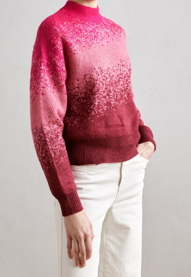 Personne portant un pull en tricot à manches longues avec un motif dégradé rose à bordeaux et un pantalon blanc, une main dans la poche, sur un fond uni.