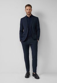 Costume tailleur navy avec un blazer à un bouton, un pantalon assorti et une chemise sombre. Caractérisé par une coupe ajustée et un tissu lisse ; chaussures en cuir.