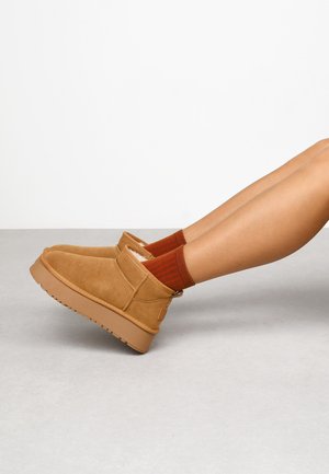 Botas de tobillo de ante marrón con forro de felpa, suela de goma y acentos cosidos. Combinadas con calcetines a rayas rojos y naranjas. Textura suave.