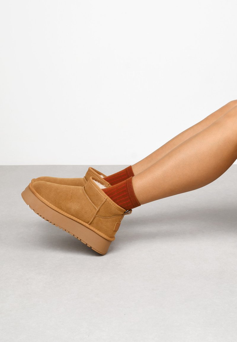 Botas de tobillo de ante marrón con forro de felpa, suela de goma y acentos cosidos. Combinadas con calcetines a rayas rojos y naranjas. Textura suave.