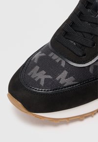 Detailný záber na prednú časť čiernych tenisiek s vzorom loga MK na sieťovinovom zvršku, čiernymi šnúrkami a gumenou textúrovanou podrážkou v hnedej farbe.