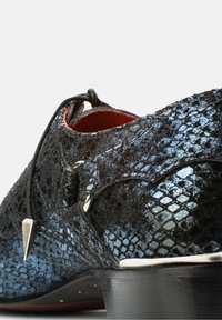 Zwarte leren schoen met textuur en metallic blauwe accenten, met een spitse neus, decoratieve hardware en een lage zwarte hak.