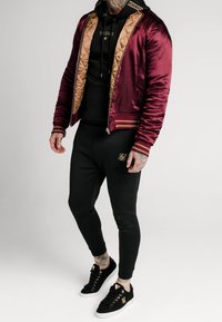 Herrebomberjakke i burgunder satin med guldquiltet for, parret med en sort hoodie, sorte tætsiddende joggers og sorte sneakers med guldakcenter.