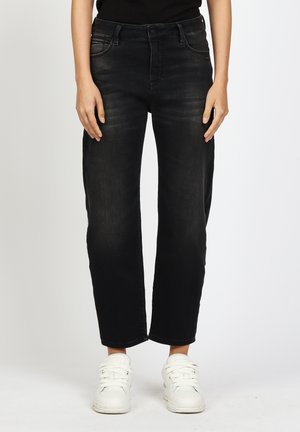 Unterkörper einer Person, die schwarze Jeans mit geradem Bein und weiße Turnschuhe trägt, vor einem einfarbigen, hellen Hintergrund stehend.