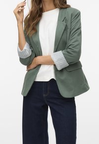 Femme portant un blazer vert retroussé sur une chemise blanche et un jean bleu foncé, posant avec un bras croisé et la main près du visage.