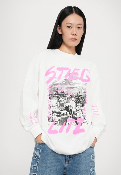 White oversized sweatshirt featuring a black and white graphic print, pink text, and relaxed fit. Paired with light blue denim jeans.