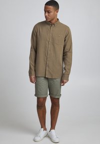 Chemise boutonnée marron clair avec une poche, portée avec un short olive retroussé et des baskets blanches, mettant en avant un tissu texturé.