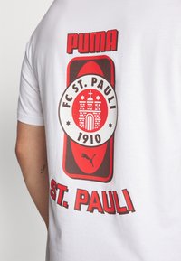 Balta kokvilnas T-krekls ar lielu FC St. Pauli logotipu, kas rotāts ar sarkaniem un brūniem akcentiem, uz kura ir rakstīts "PUMA" un "1910" treknā tekstā.