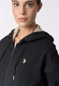 Felpa nera con zip e cappuccio, con interno beige, caratterizzata da un piccolo logo ricamato sul petto, realizzata in tessuto morbido con una superficie liscia.