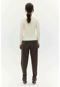 Lichtbeige, geribde trui met een getailleerde silhouette en gouden schouderaccenten, gecombineerd met losse, bruine broek en hakken.