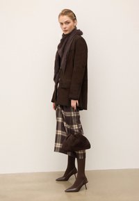 Veste en daim marron, écharpe duveteuse violette foncée, jupe à carreaux dans des tons de marron et de crème, sac à main rond marron, talons pointus marron, texture lisse.
