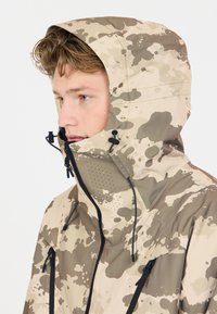 Beige ja brun camuflaj jakke med høy krage, justerbar hette, glidelåser og perforerte detaljer for ventilasjon.