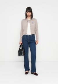 Beige gebreide cardigan met knoopsluiting, blauwe borduursels, witte top, blauwe rechte spijkerbroek en zwarte tas. Glanzende bordeauxrode schoenen.