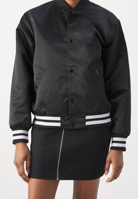 Schwarze Bomberjacke mit glatter, glänzender Oberfläche, ausgestattet mit einem Stehkragen, vorne mit Knöpfen und weißen Streifen an den Bündchen und dem Saum.