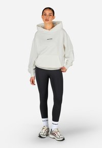 Sudadera blanca con bolsillo frontal y logo negro, combinada con leggings negros, calcetines blancos y zapatillas deportivas plateadas. Tejido suave y corte relajado.