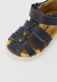 Tummansininen nahkasandaali, jossa on useita hihnoja, keltaisia yksityiskohtia ja pehmustettu beige pohja. Varustettu kumipohjalla, joka tarjoaa pitoa.