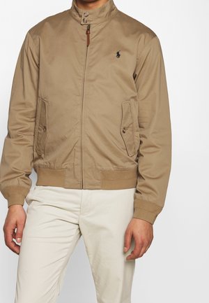 Mężczyzna ubrany w beżową kurtkę na zamek błyskawiczny z kołnierzem zapinanym na guziki oraz kremowe spodnie, stoi z rękami swobodnie opuszczonymi wzdłuż ciała.