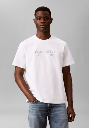 Mann trägt ein weißes Calvin Klein T-Shirt mit dem Schriftzug „Calvin Klein EST. 1968“, kombiniert mit blauen Jeans, steht vor grauem Hintergrund.