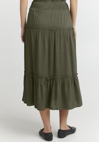Gonna maxi di colore verde oliva con volant a strati, con una vita arricciata e orlo arruffato, realizzata in tessuto leggero. Indossata con scarpe basse nere.