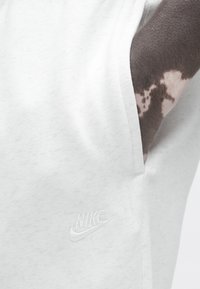 Baltos Nike sportinės kelnės su lygiu paviršiumi turi kairiąją kišenę ir siuvinėtu logotipu tokios pačios spalvos. Audinyje subtilus raštas.