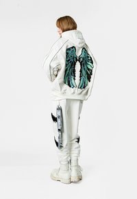 Chaqueta bomber blanca con bordado de alas verdes y negras en la parte posterior, combinada con joggers blancos que tienen acentos negros y una correa de utilidad.