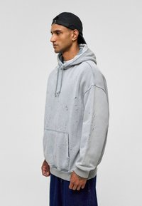 Lichtgrijze oversized hoodie met een voorzak, voorzien van een trekkoordcapuchon en gespetterde zwarte accenten. Zacht materiaal met ribgebreide mouwen.