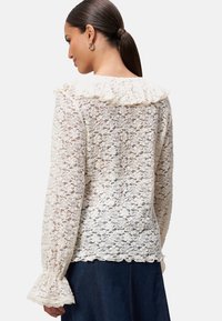 Kantblouse in gebroken wit met een gerimpelde kraag en klokmouwen, voorzien van ingewikkelde bloemmotieven en een golfvormige zoom.