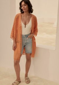 Orange spetskimono med blommönster över en vit body, matchad med högmidjade denimshorts och beige slidesandaler.