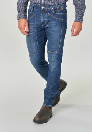 Jeans in denim blu con una vestibilità slim, caratterizzati da cuciture a contrasto, tasche frontali con un design curvo unico, abbinati a stivali marroni.