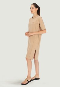 Beżowa sukienka do kolan z krótkimi rękawami, okrągłym dekoltem, bocznymi rozcięciami i gładką teksturą, w połączeniu z płaskimi czarnymi sandałami z złotymi akcentami.