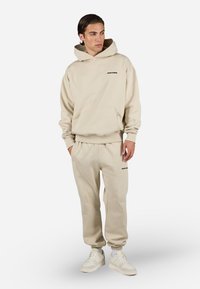Beige hettegenser og matchende joggebukser laget av mykt stoff. Har kengurulomme, ribbestrikkede mansjetter og svarte logoakenter.