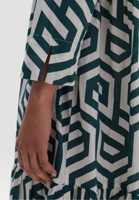 Grön och vit mönstrad klänning med geometrisk design, med trekvartslånga ärmar, sidoflikar och en rynkad fåll.