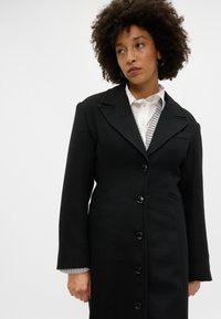 Černý tailored kabát s výstřihem do V, šesti knoflíky a dvěma bočními kapsami. Pod ním je vidět světle šedý pruhovaný svetr.