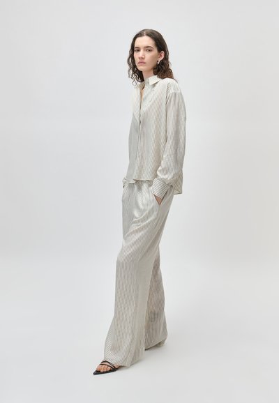 Nő áll, kezét zsebre téve, laza, világos csíkos ingben és hozzáillő, bőszárú nadrágban, fekete nyitott orrú magas sarkú cipővel.