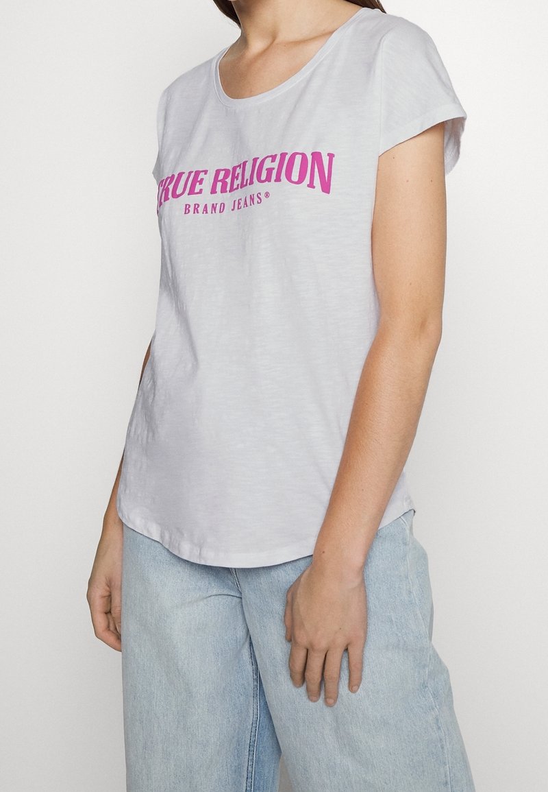 Světle šedé tričko s krátkým rukávem vyrobené z měkké látky, které má výrazné růžové logo "TRUE RELIGION" přes hruď. Uvolněný střih a zaoblený lem.