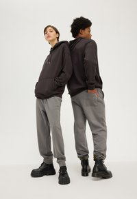Deux individus se tiennent dos à dos, vêtus de sweats à capuche sombres et de pantalons de survêtement gris ; tous deux portent des bottes à plateforme noires. Le tissu est doux et lisse.