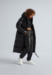 Parka negra larga con capucha, textura brillante y diseño acolchado. Combinada con pantalones negros ajustados y zapatillas blancas.