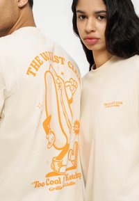 Άνδρας και γυναίκα φορούν μπεζ μπλούζες με πορτοκαλί κείμενο· η μπλούζα του άνδρα δείχνει καρτούν χοτ ντογκ και το λογότυπο "The Wurst Club" στην πλάτη, της γυναίκας στο μπροστινό μέρος.