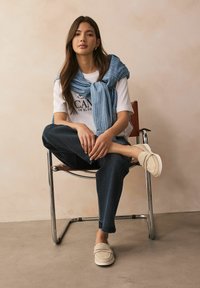 White t-shirt with graphic logo, light blue striped scarf draped over shoulders, dark jeans, and beige slip-on shoes on a metal chair.