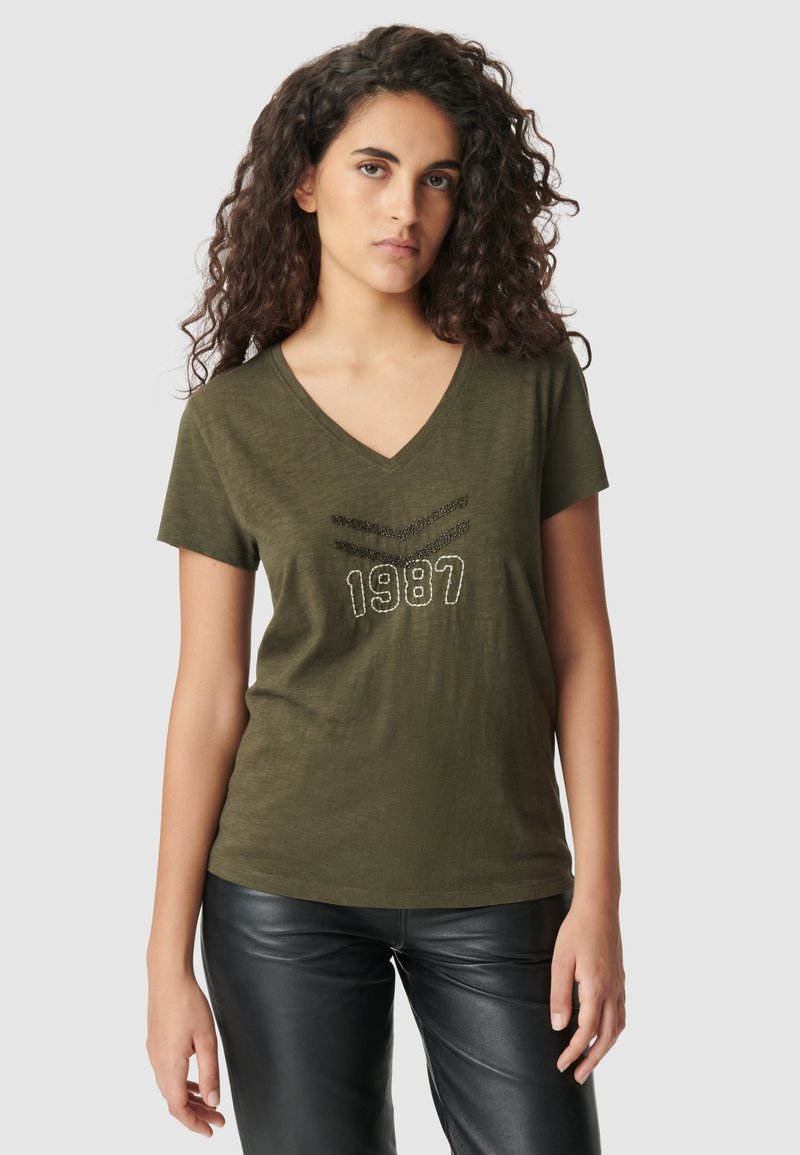 Olijfgroene v-hals t-shirt met korte mouwen, met een gestructureerde graphic "1987" en versieringen aan de voorkant. Draag het met zwarte leren broek.