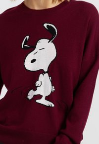 Pull en tricot bordeaux avec un grand design de Snoopy noir et blanc, col rond et coupe décontractée. Texture douce et manches longues.