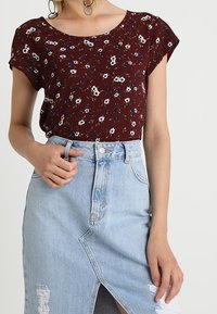 Bordeauxfarbener, kurzärmeliger Oberteil mit floralem Muster, kombiniert mit einem hellblauen, verwaschenen Jeansrock mit einem Schlitz vorne und Knopfverschluss.