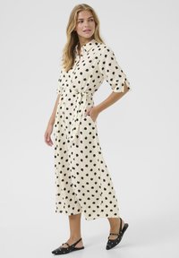 Abito a pois in crema con macchie nere, caratterizzato da una chiusura con bottoni sul davanti, colletto, maniche corte e una cintura da annodare. Include tasche.