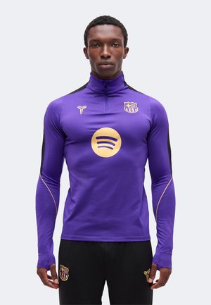 Homme portant une veste de sport violette ajustée avec un col montant affichant les logos de Barcelone et Spotify, associée à un pantalon noir.