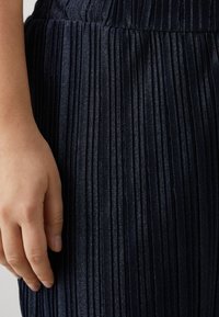 Navy pleated skirt with a smooth, shiny texture. Features narrow, vertical folds and a waistband, partially showing a hand resting on it.