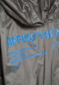 Szary materiał z niebieskim napisem "OFF DUTY GEAR", "W-F i TRENING" oraz "WYSOKOGÓRSKIE NISKIE OTWARCIE" na kurtce lub torbie.