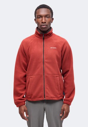 Homme debout face à l'avant portant une veste polaire Columbia rouge rouille avec poches avant et fermeture éclair, pantalon gris visible en dessous.