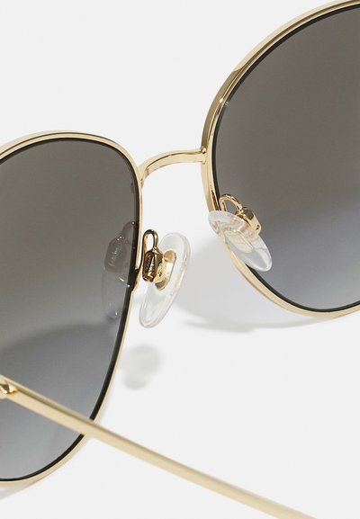 Ochelari de soare din metal auriu cu lentile gri închis. Prezintă pernuțe nazale transparente și o formă alungită, rotundă. Design elegant, minimalist.
