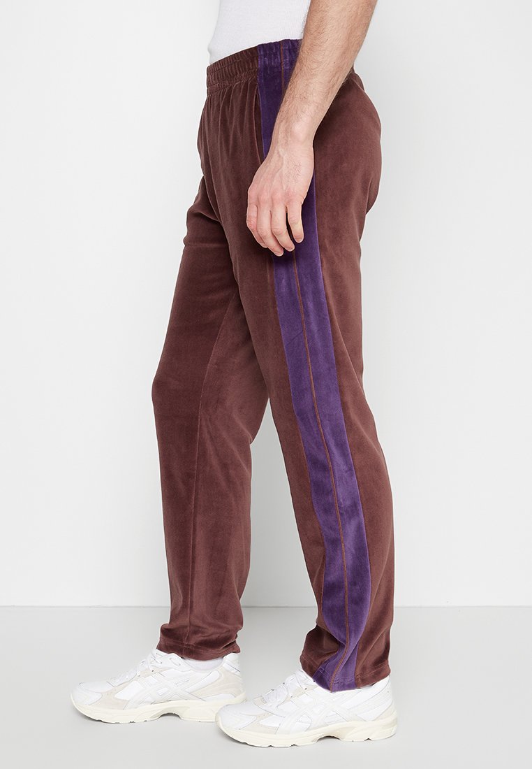 Pantalons de jogging en velours marron avec une bande latérale violette, dotés d'une taille élastique et d'un design de jambe fuselée, associés à des baskets blanches.